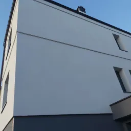 Nowoczesny budynek wielorodzinny z jasną elewacją i ciemną podmurówką, nietypowe, wąskie okna, widok z dołu na błękitne niebo.