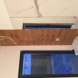 Fragment łazienki z drewnopodobną zabudową nad oknem, białą armaturą i marmurowymi płytkami na ścianie, widoczne kable wystające z zabudowy.