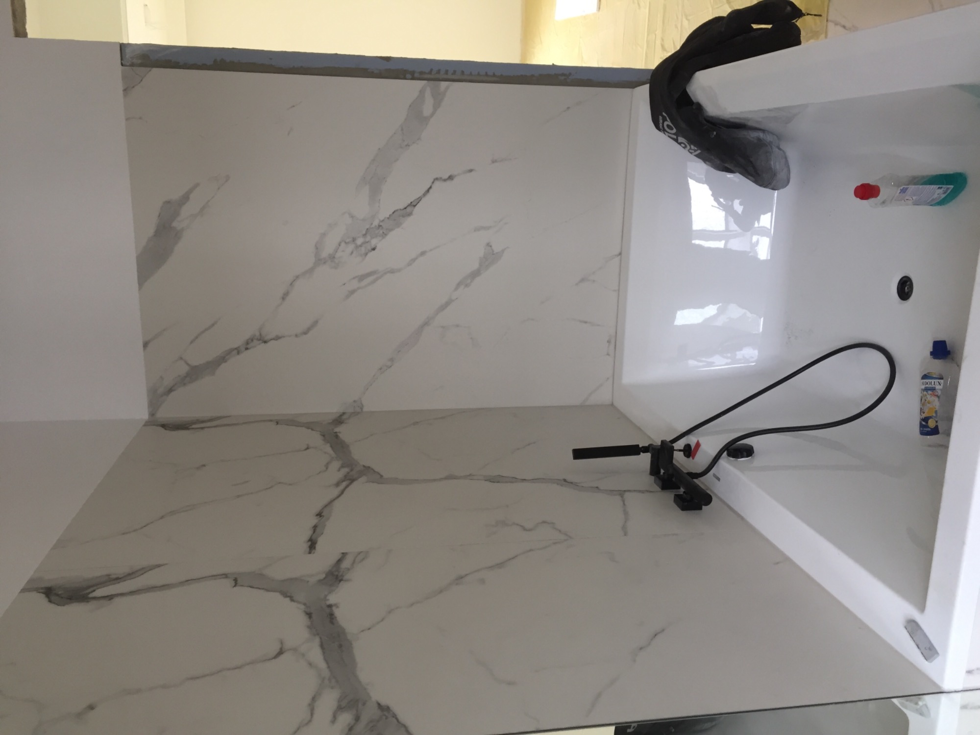 Białe płytki imitujące marmur na ścianie i podłodze w trakcie układania, widoczny fragment wanny i narzędzie do cięcia płytek.