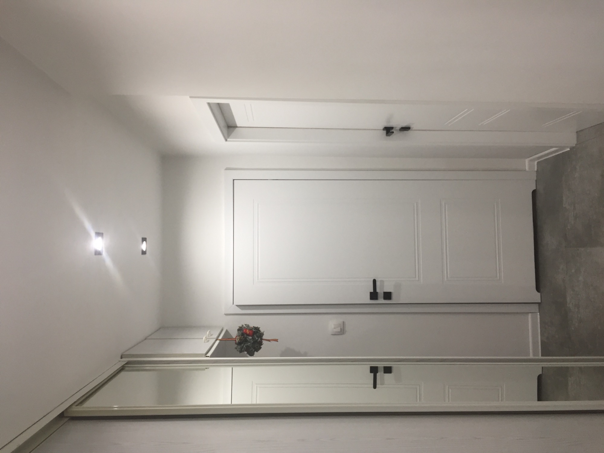 Białe, dwuskrzydłowe drzwi wewnętrzne z czarnymi klamkami, widoczne w przedpokoju z szafą wnękową z lustrem i dekoracyjnym wieńcem.