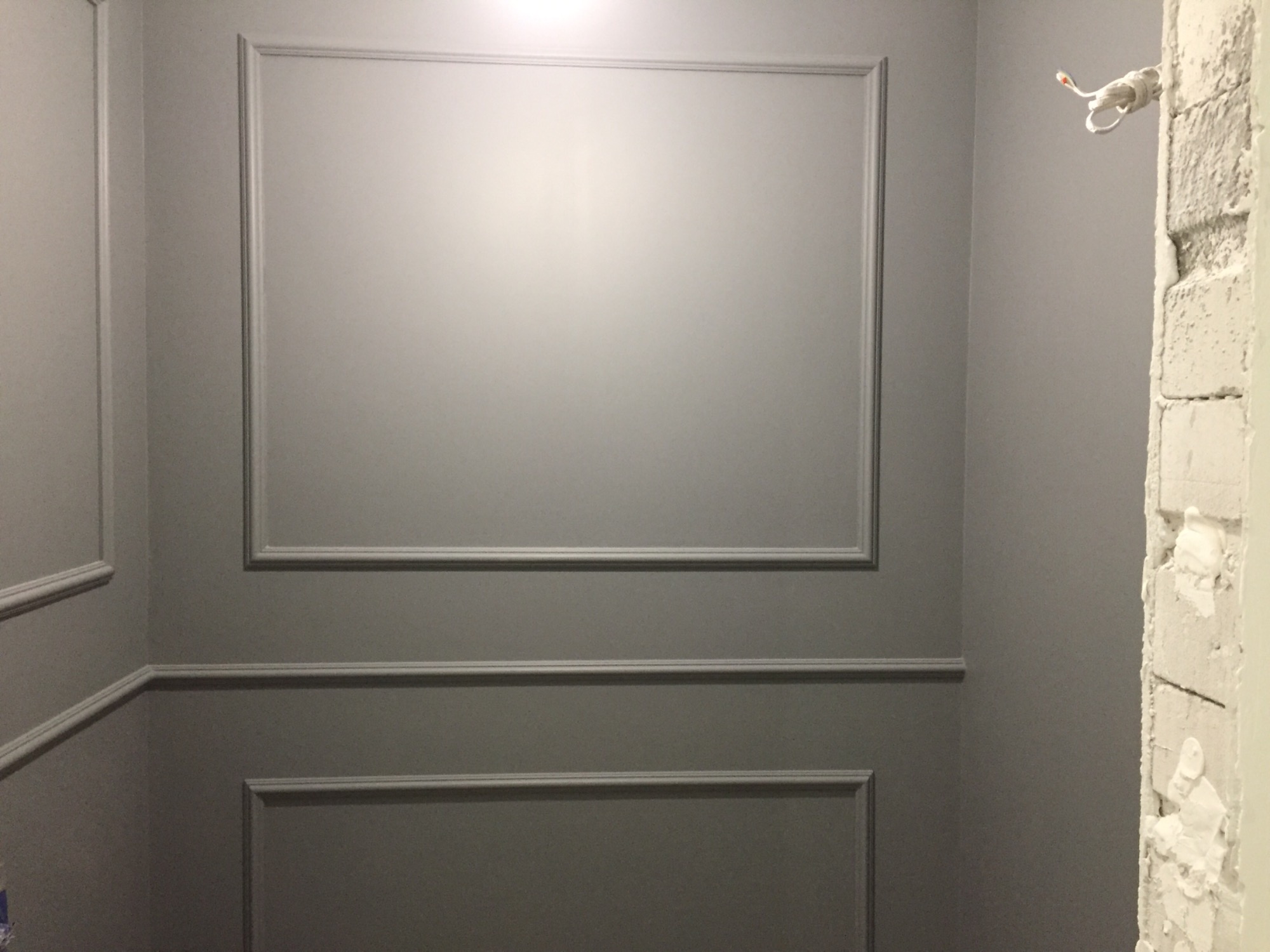 Ściana pomalowana na szaro z dekoracyjnymi listwami w stylu klasycznym; widoczny fragment surowego muru z wystającymi przewodami elektrycznymi.