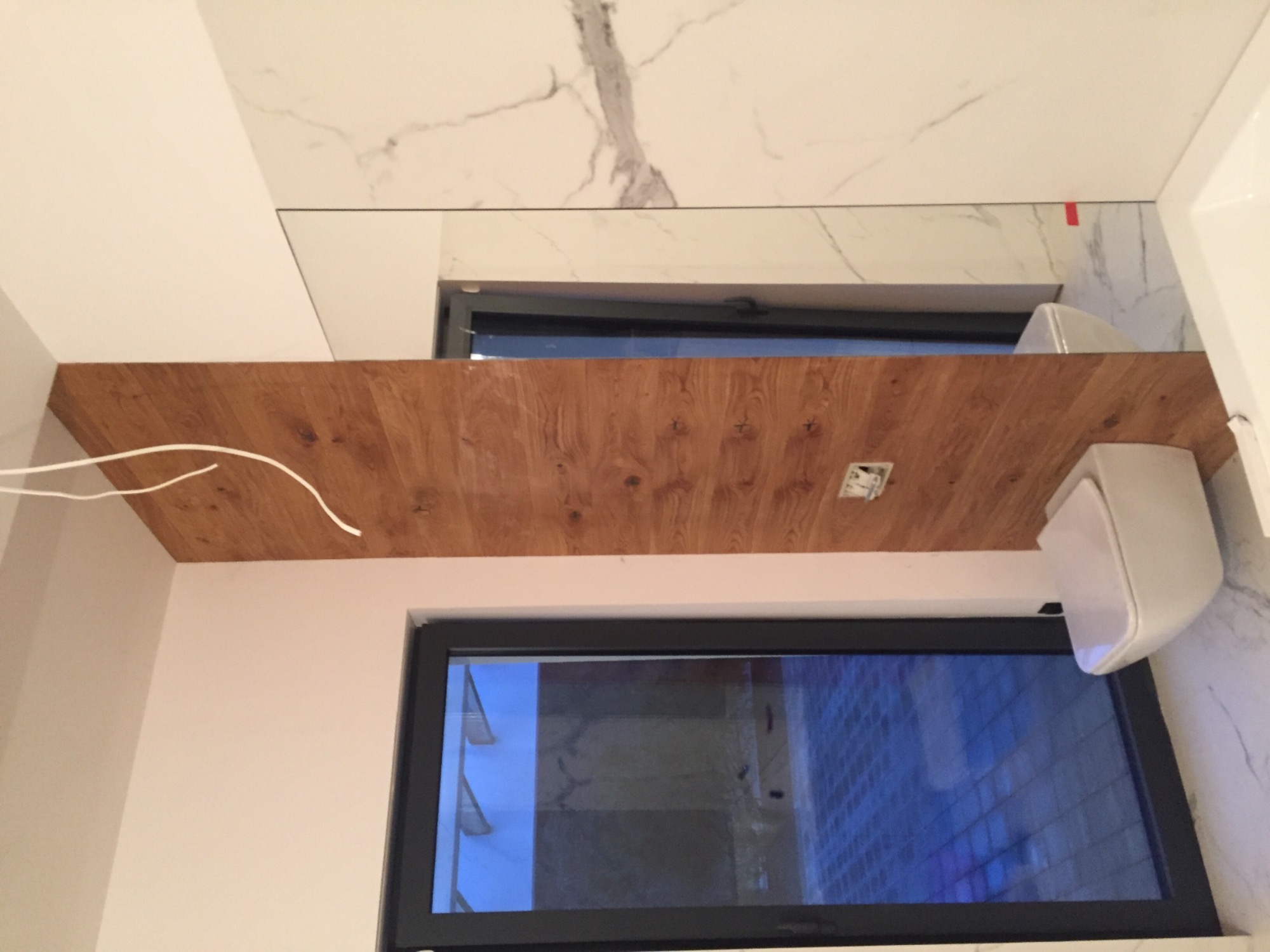 Fragment łazienki z drewnopodobną zabudową nad oknem, białą armaturą i marmurowymi płytkami na ścianie, widoczne kable wystające z zabudowy.