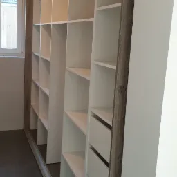 Biała szafa wnękowa z szufladami i półkami, obudowana drewnopodobną płytą, widoczna na tle szarej podłogi i okna.