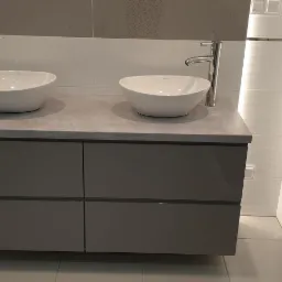 Nowoczesna łazienka z podwójną umywalką nablatową na szarym blacie i szafką w kolorze gołębim, oświetlenie LED wzdłuż ściany z białych płytek.