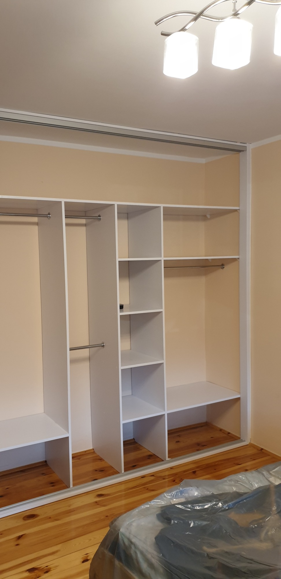 Biała, otwarta szafa wnękowa z półkami i drążkami na ubrania, zamontowana w pokoju z drewnianą podłogą, widoczna część mebla przykryta folią ochronną.