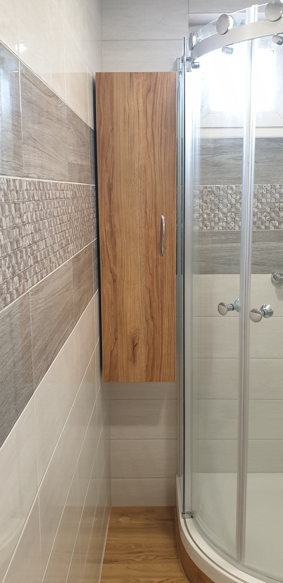 Wąska, drewniana szafka wisząca obok kabiny prysznicowej w łazience w Zieminie, z widocznymi płytkami ceramicznymi na ścianach i panelami podłogowymi.