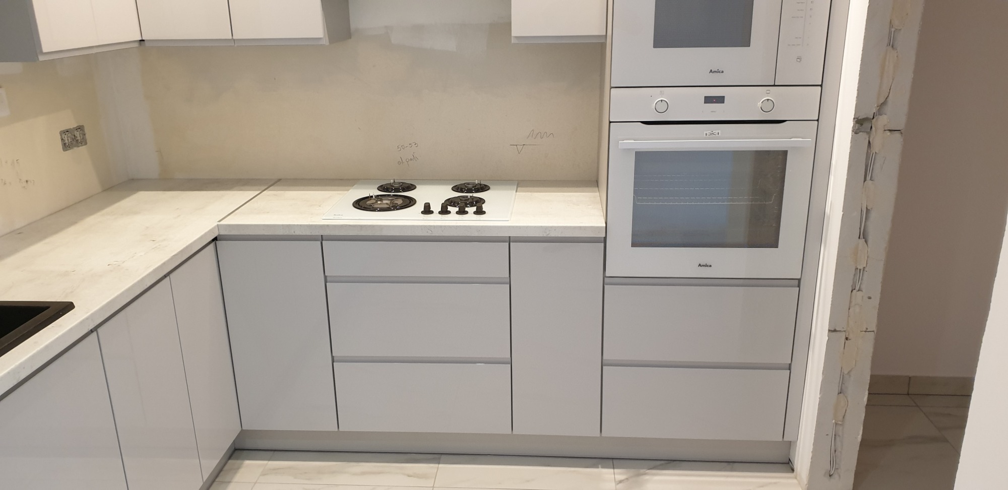 Jasnoszara zabudowa kuchenna z białym blatem, płytą grzewczą i piekarnikiem Amica, widoczne ślady po pracach wykończeniowych na ścianie.