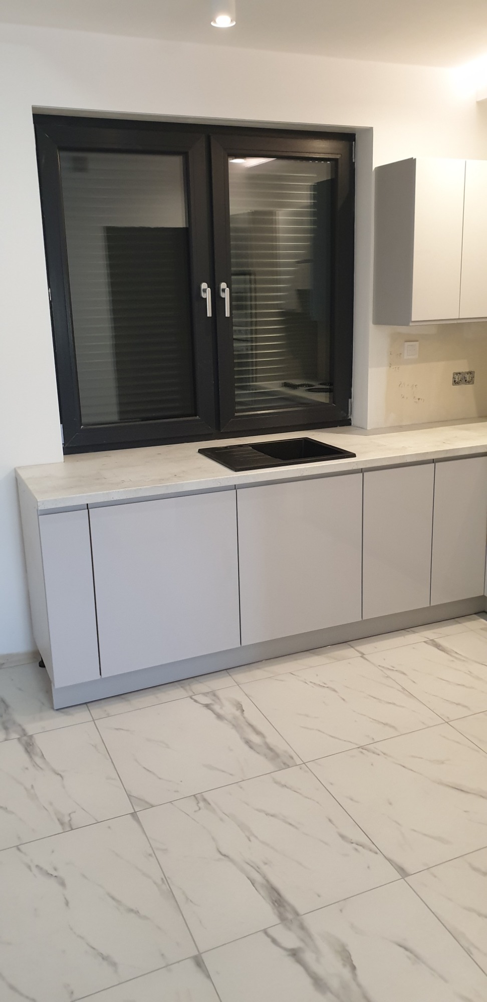 Szare, minimalistyczne szafki kuchenne z blatem imitującym marmur, czarny zlew pod oknem z żaluzjami, płytki podłogowe imitujące marmur.