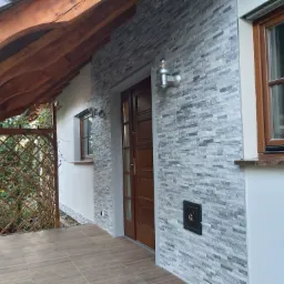 Elewacja domu wykończona szarą, warstwową okładziną kamienną, oświetlona srebrnymi kinkietami z żarówkami w stylu retro, z drewnianą stolarką okienną i zadaszeniem tarasu z belek