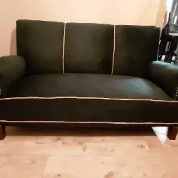 Ciemnozielona sofa dwuosobowa z ozdobnymi gwoździami i wypustkami, widoczna z przodu na tle jasnej ściany i podłogi.