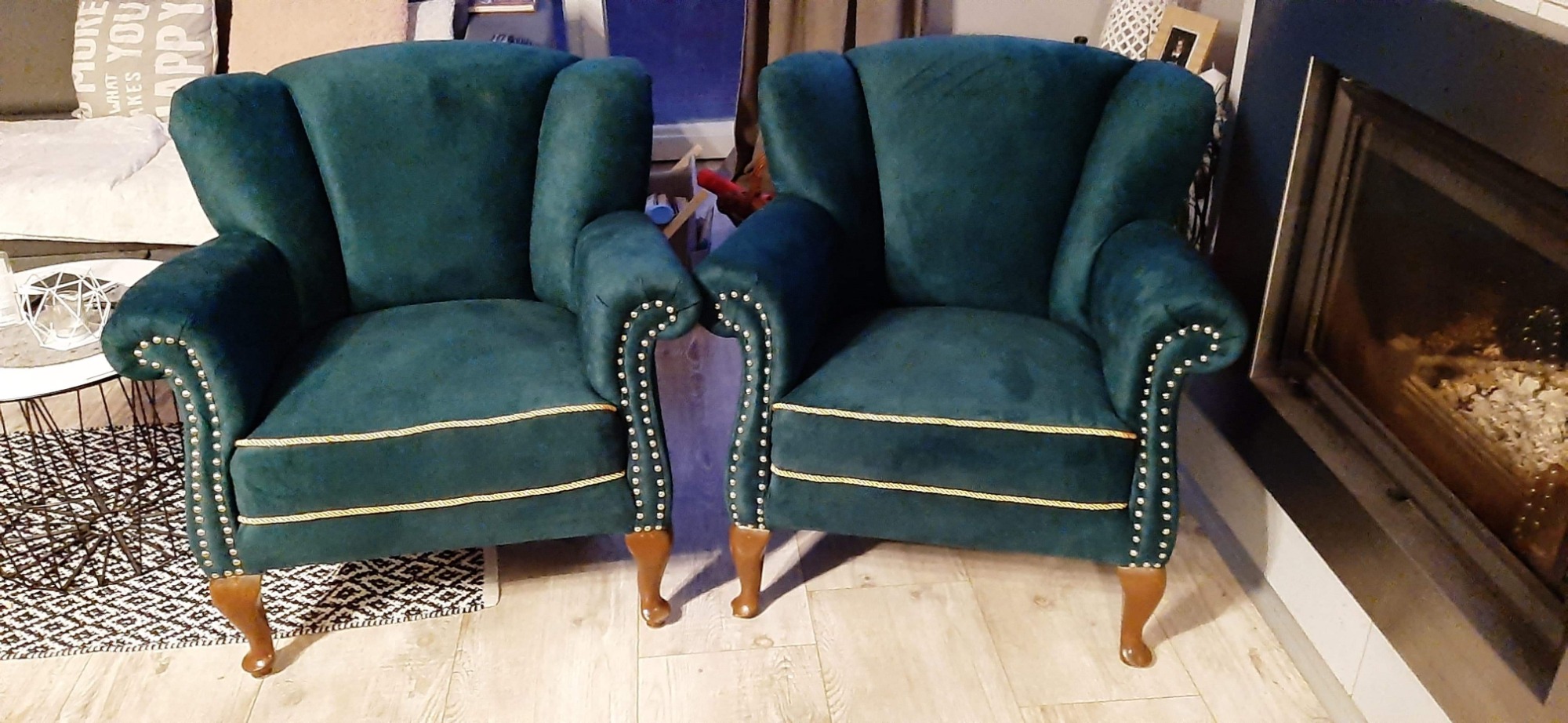 Dwa identyczne, szmaragdowe fotele typu uszak z drewnianymi nóżkami i ozdobnymi gwoździami tapicerskimi, stojące na jasnej podłodze obok kominka.