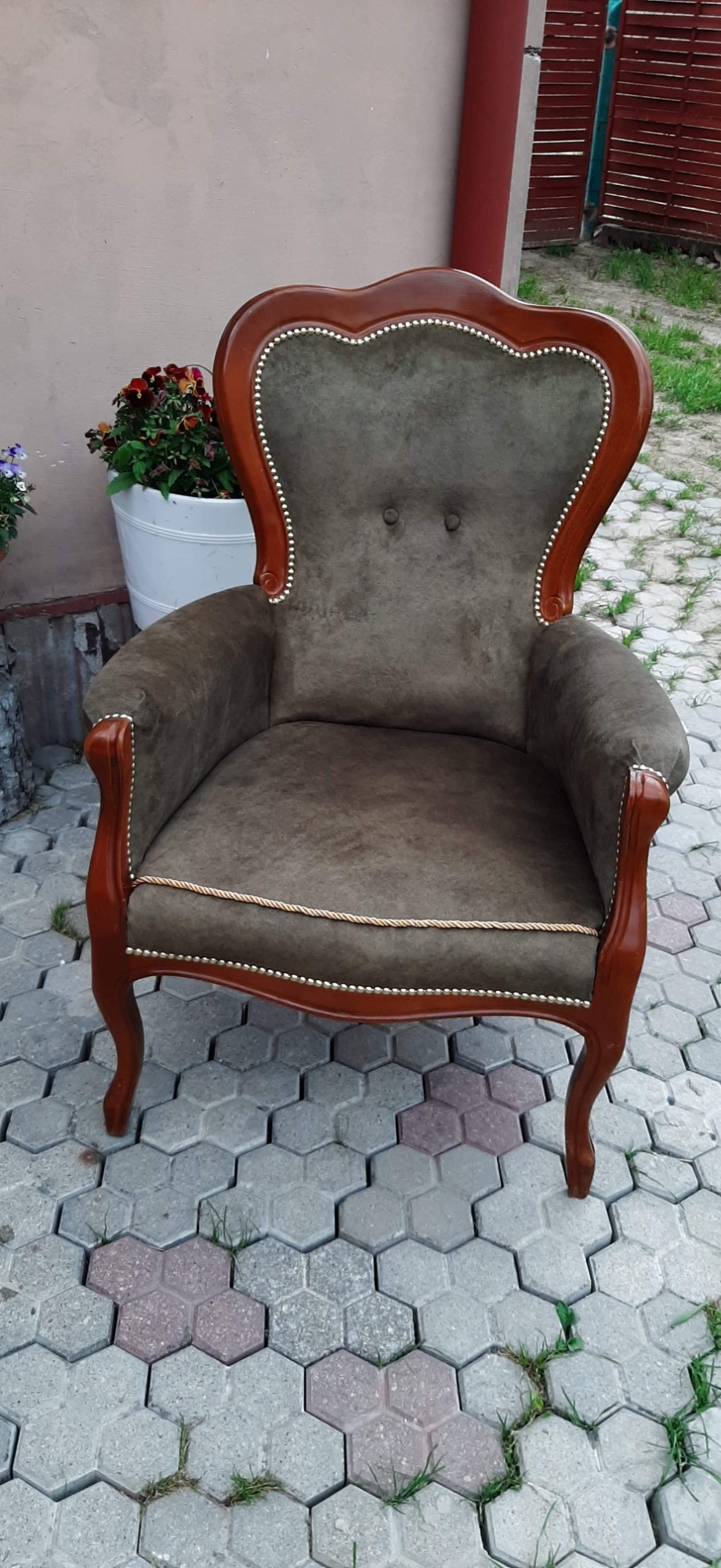 Tapicerowany fotel typu uszak w stylu retro z drewnianymi, giętymi nogami i dekoracyjnymi gwoździami, stojący na szarej kostce brukowej obok donicy z kwiatami.