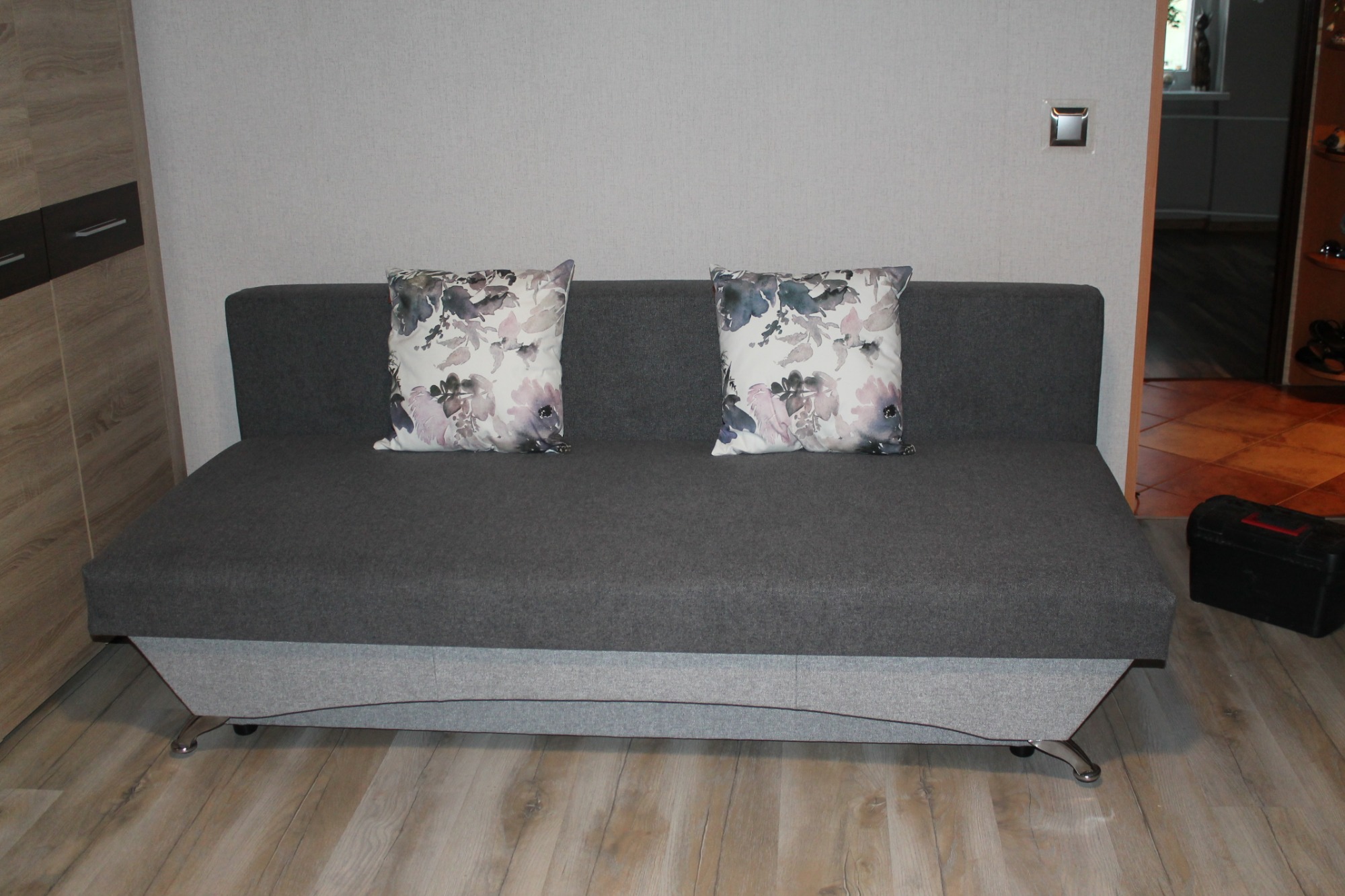 Rozłożona szara sofa z dwoma poduszkami w kwiaty, chromowane nóżki, widoczna szafa i płytki podłogowe imitujące drewno.