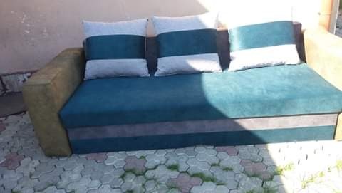 Rozkładana sofa z turkusową tapicerką i poduszkami w paski, ustawiona na zewnątrz na tle kostki brukowej.