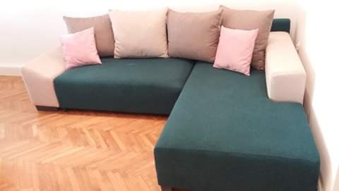 Narożnik z ciemnozieloną tapicerką i beżowymi bokami, ozdobiony poduszkami w odcieniach różu i beżu, ustawiony na parkiecie w jodełkę.