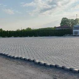 Rozległy, szary plac z kostki brukowej typu 'I', obrzeża z kostki, w tle szpaler tuj, budynek z ciemnym dachem, ciężarówka Volvo i dwa busy.