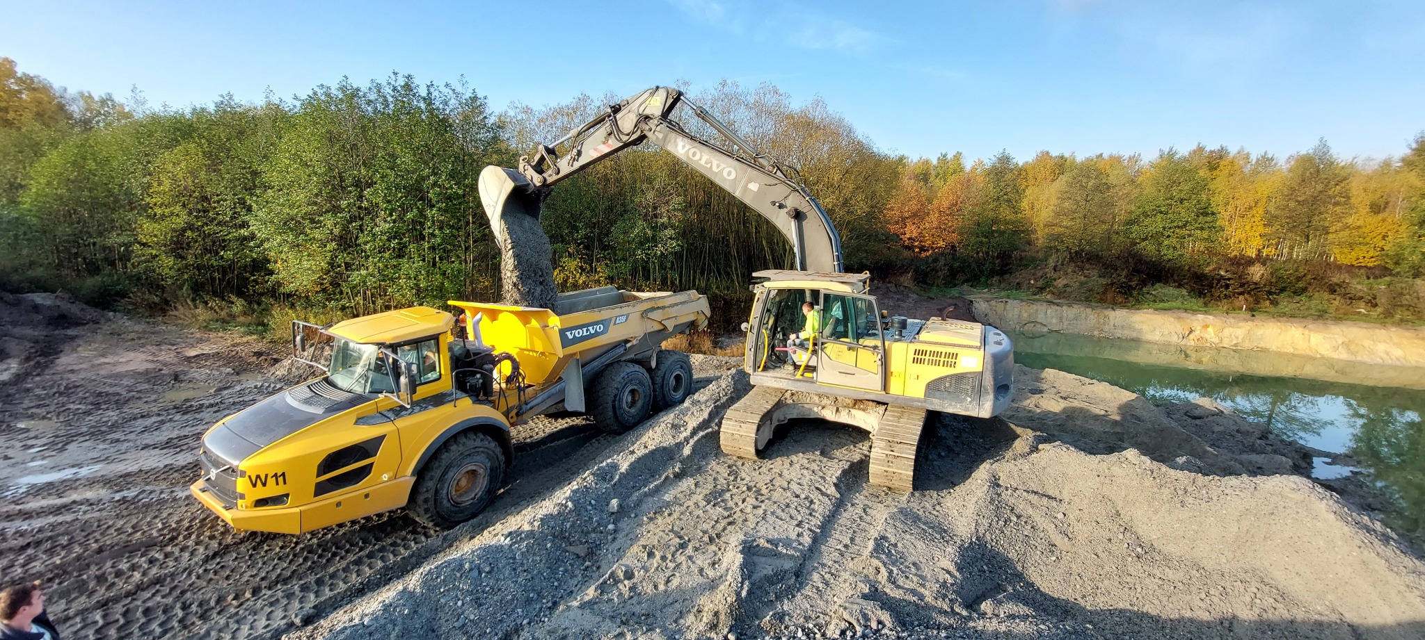 Żółty wywrotka Volvo A25F odbiera urobek z łyżki koparki Volvo na tle wyrobiska z wodą i drzew.
