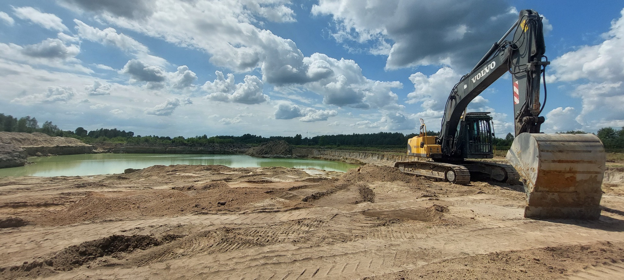 Żółto-czarna koparka Volvo na terenie budowy, z widocznym wyrobiskiem z wodą w tle, pod błękitnym niebem z obłokami.