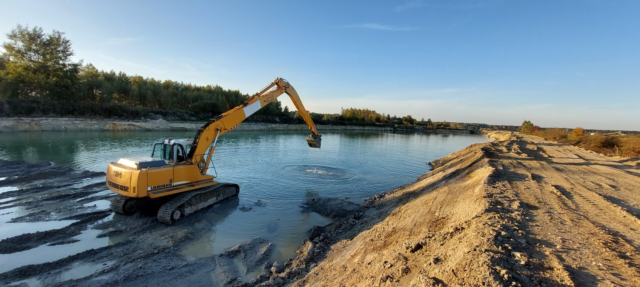 Żółta koparka Liebherr na brzegu zbiornika wodnego, ramię z łyżką zanurzone w wodzie, widoczne ślady po kołach w błocie, drzewa w tle.