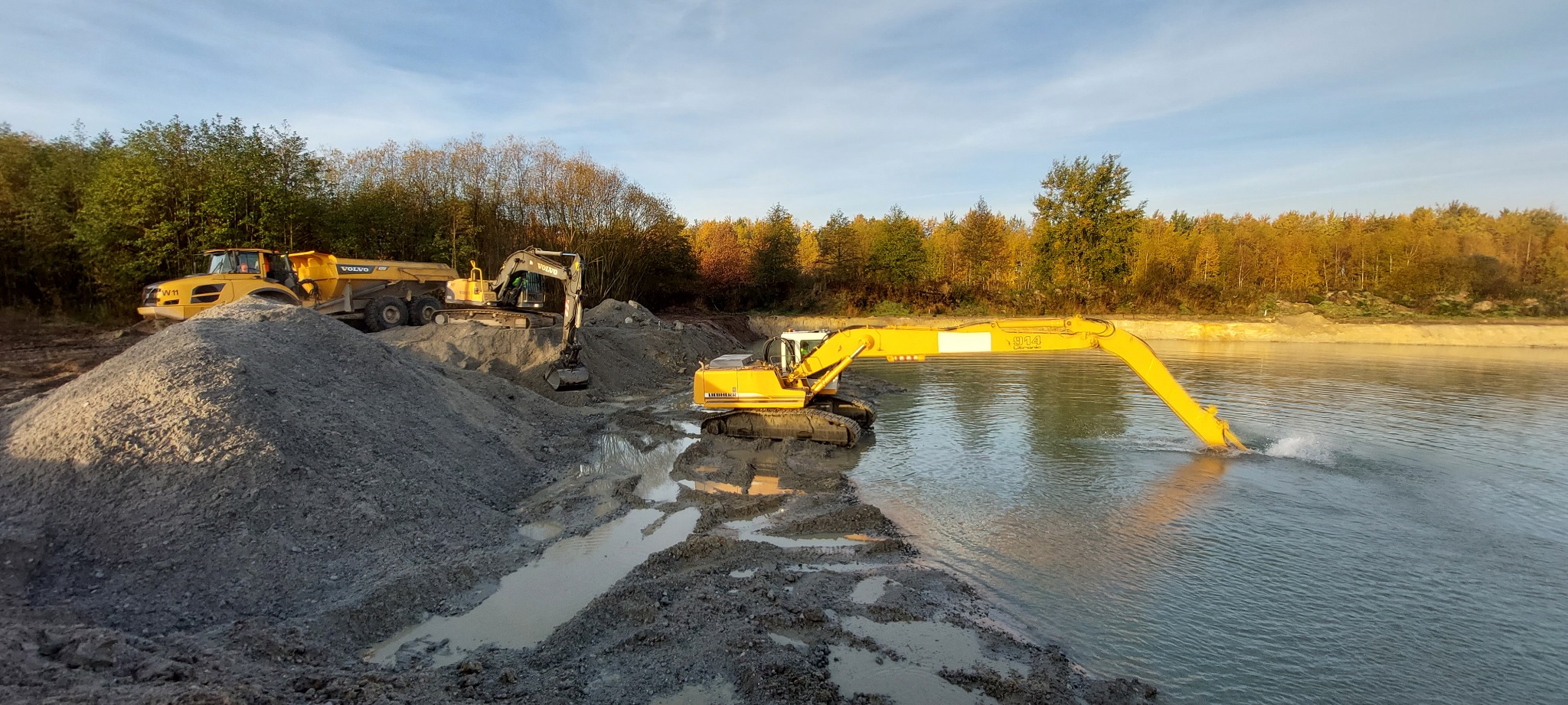 Żółta koparka Liebherr 914 pracująca w zbiorniku wodnym, obok hałda ziemi i wywrotka Volvo W11 na tle drzew.