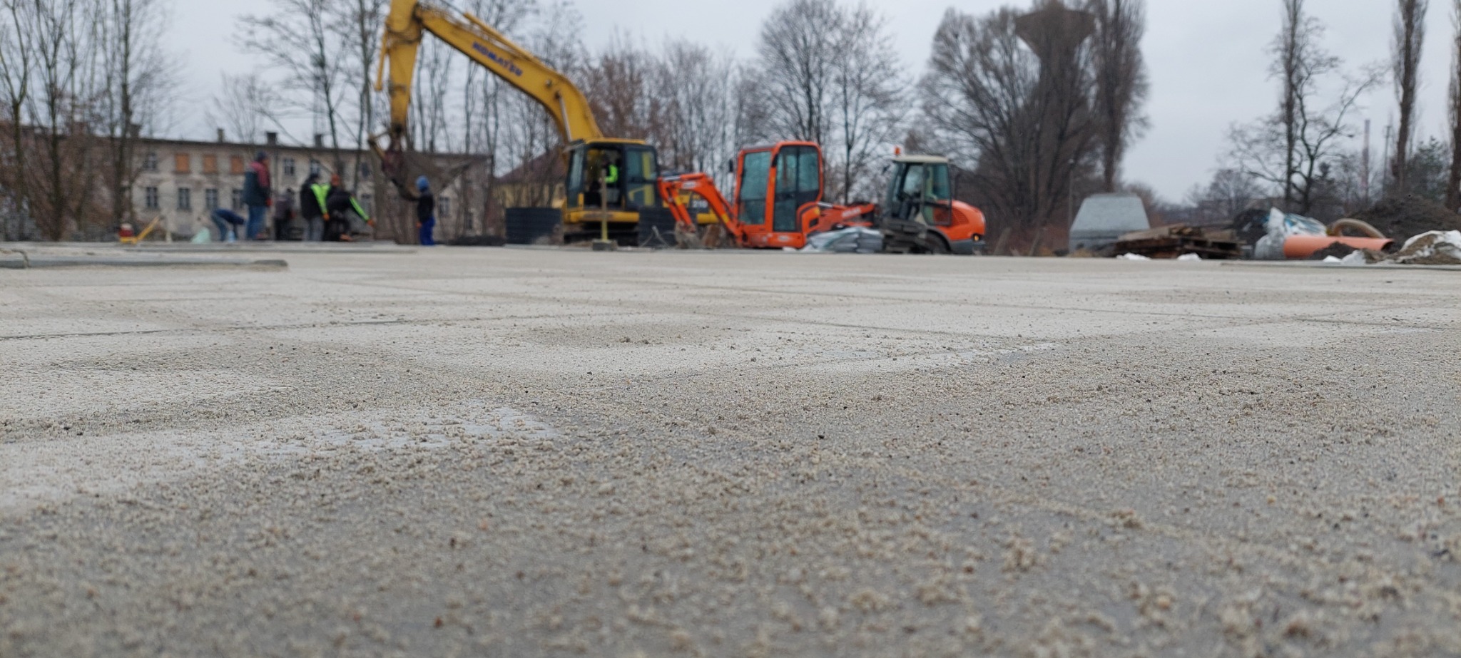 Przygotowany teren pod budowę drogi z widocznymi maszynami budowlanymi: koparka, mini koparka i walec drogowym w tle, oraz grupą robotników w oddali.
