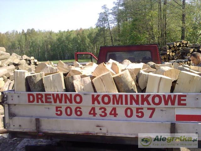 Przyczepa wypełniona pociętymi kawałkami drewna na tle lasu, z czerwonym napisem 'DREWNO KOMINKOWE' i numerem telefonu na burcie.