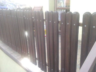 Ciemnobrązowe sztachety brzozowe tworzące ogrodzenie, mocowane na gwoździe, z widocznym fragmentem parapetu i rozproszonym światłem słonecznym w tle.