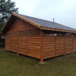 Drewniana konstrukcja z ażurowymi ścianami i dwuspadowym dachem pokrytym gontem bitumicznym, posadowiona na betonowych bloczkach z pomarańczowymi rurami, na tle trawnika i szarego samochodu.