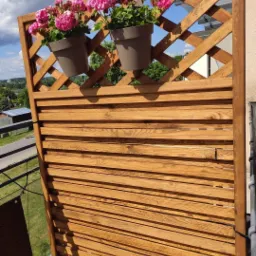 Drewniana przesłona balkonowa z kratownicą i poziomymi deskami, na której zawieszone są dwie donice z różowymi kwiatami, widok na zielony krajobraz w tle.