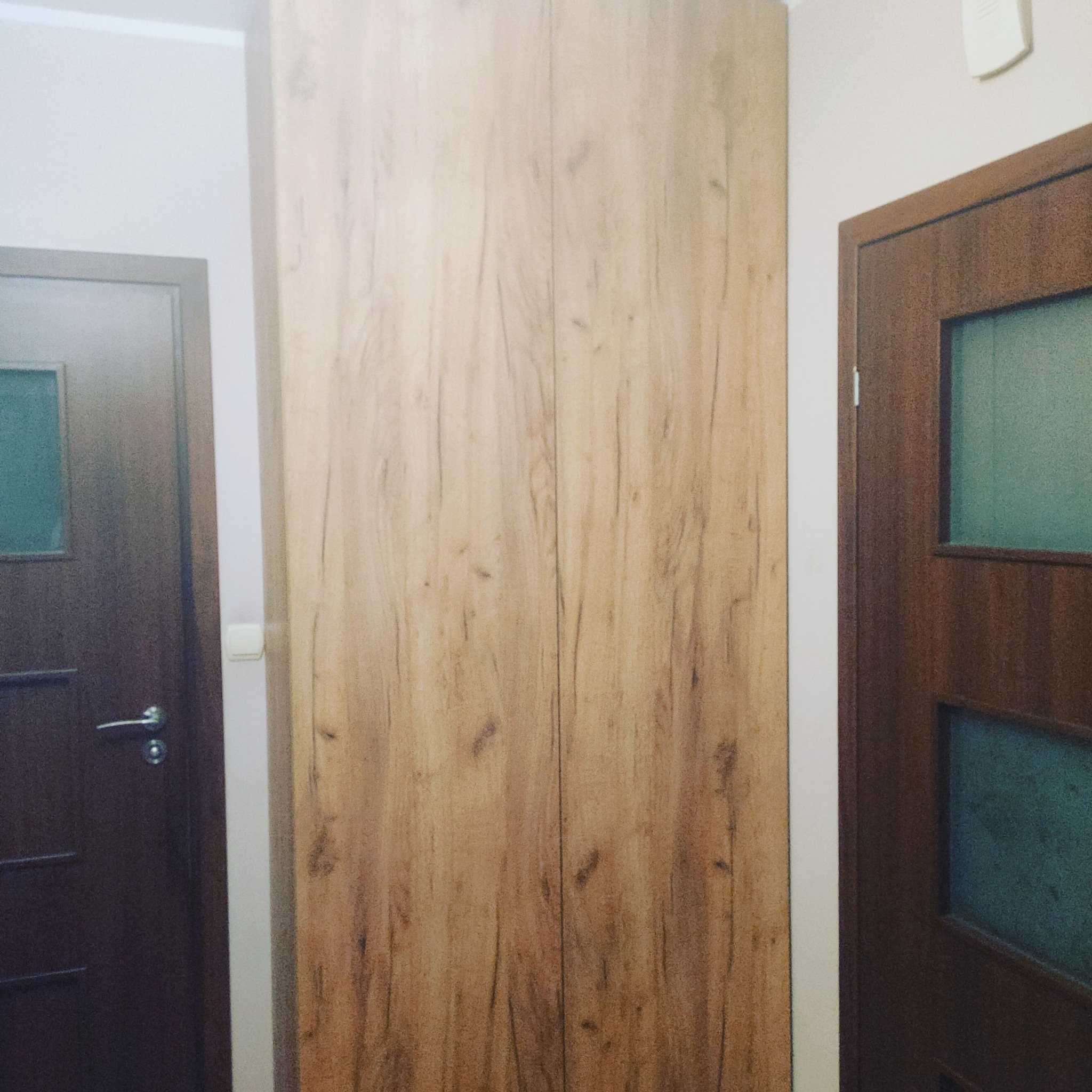 Dwudrzwiowa szafa wnękowa z jasnego drewna, wkomponowana w przestrzeń między dwoma drzwiami w korytarzu.