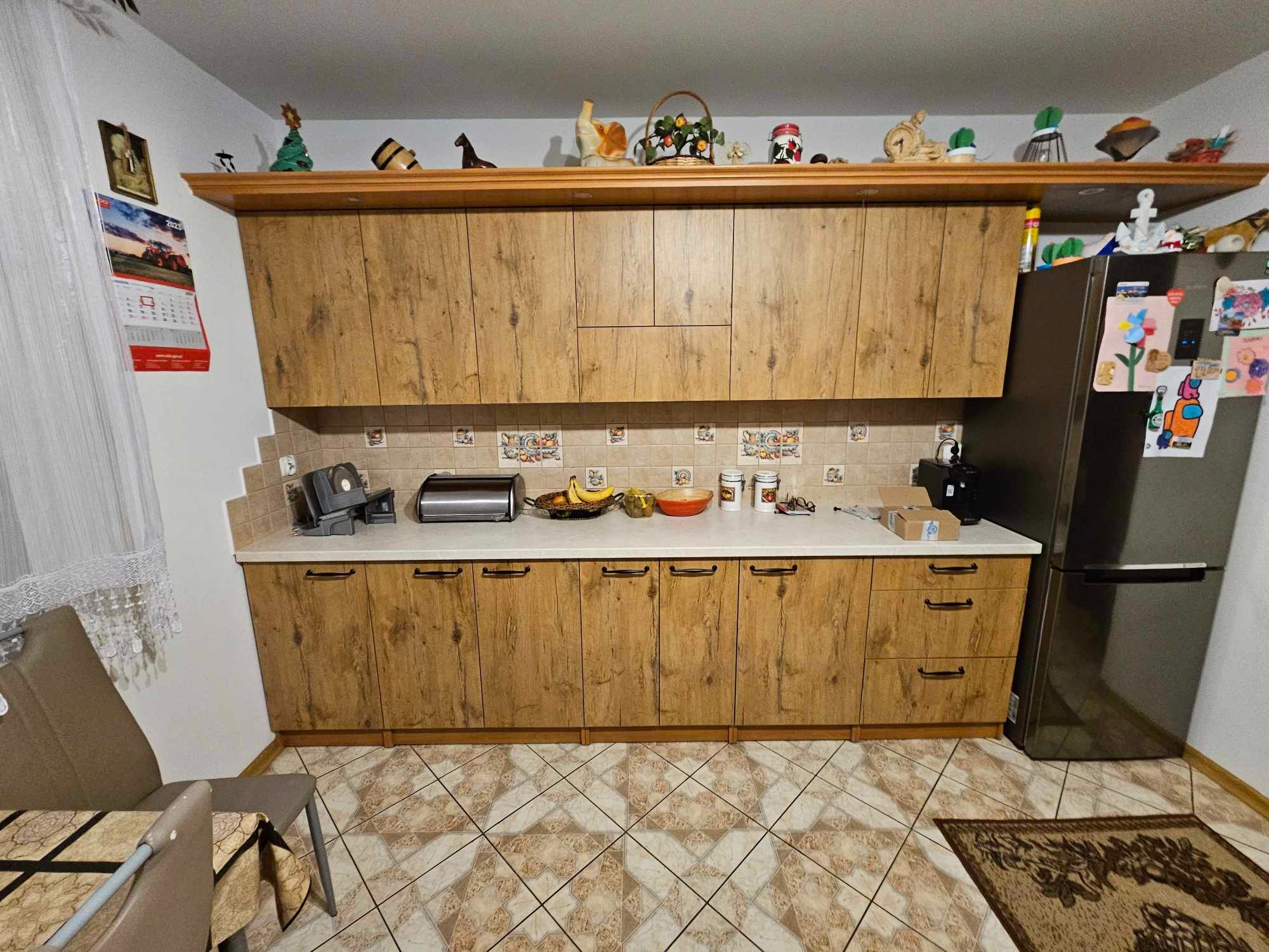 Kuchnia z drewnopodobnymi frontami szafek, biały blat, płytki na ścianie i podłodze w odcieniach beżu, dekoracje na górnej półce, widoczna lodówka i fragment stołu z krzesłami.