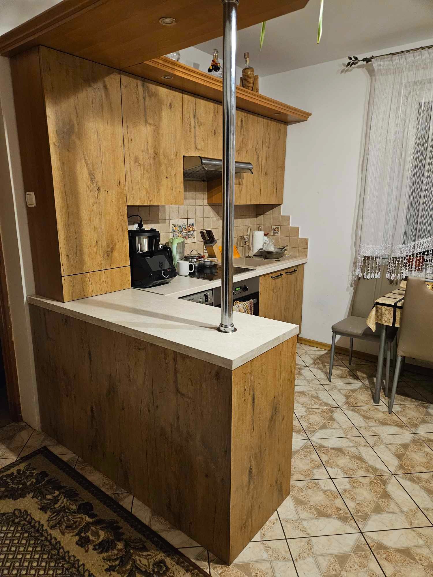 Wnętrze kuchni z drewnianymi szafkami o fakturze drewna, białym blatem i metalowym słupkiem, widoczne sprzęty AGD, stół i krzesła, płytki podłogowe o wzorze geometrycznym.