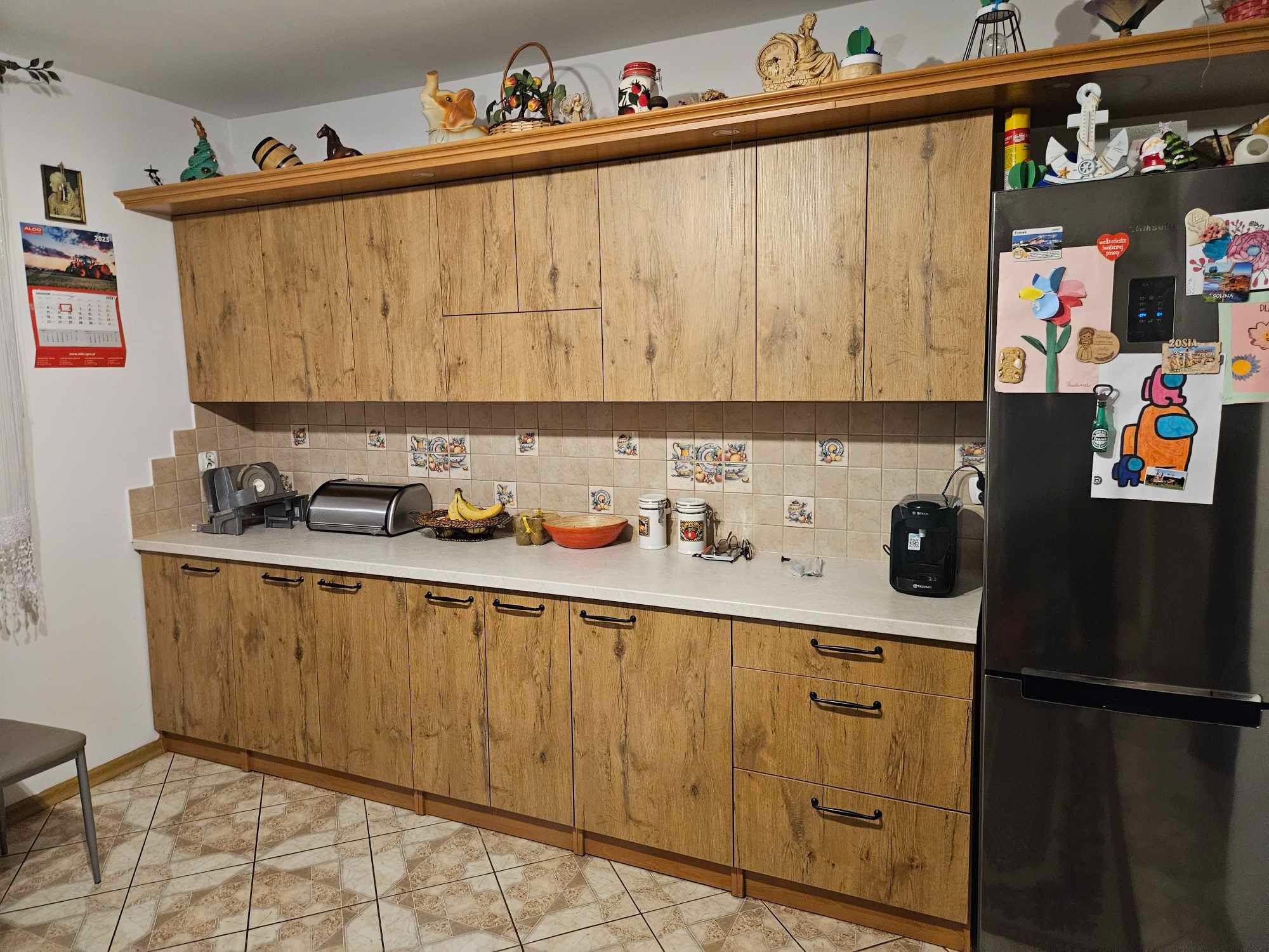 Kuchnia z drewnianymi szafkami na wymiar, jasny blat, płytki z motywem owoców i warzyw, lodówka z magnesami, różne dekoracje na górnej półce.