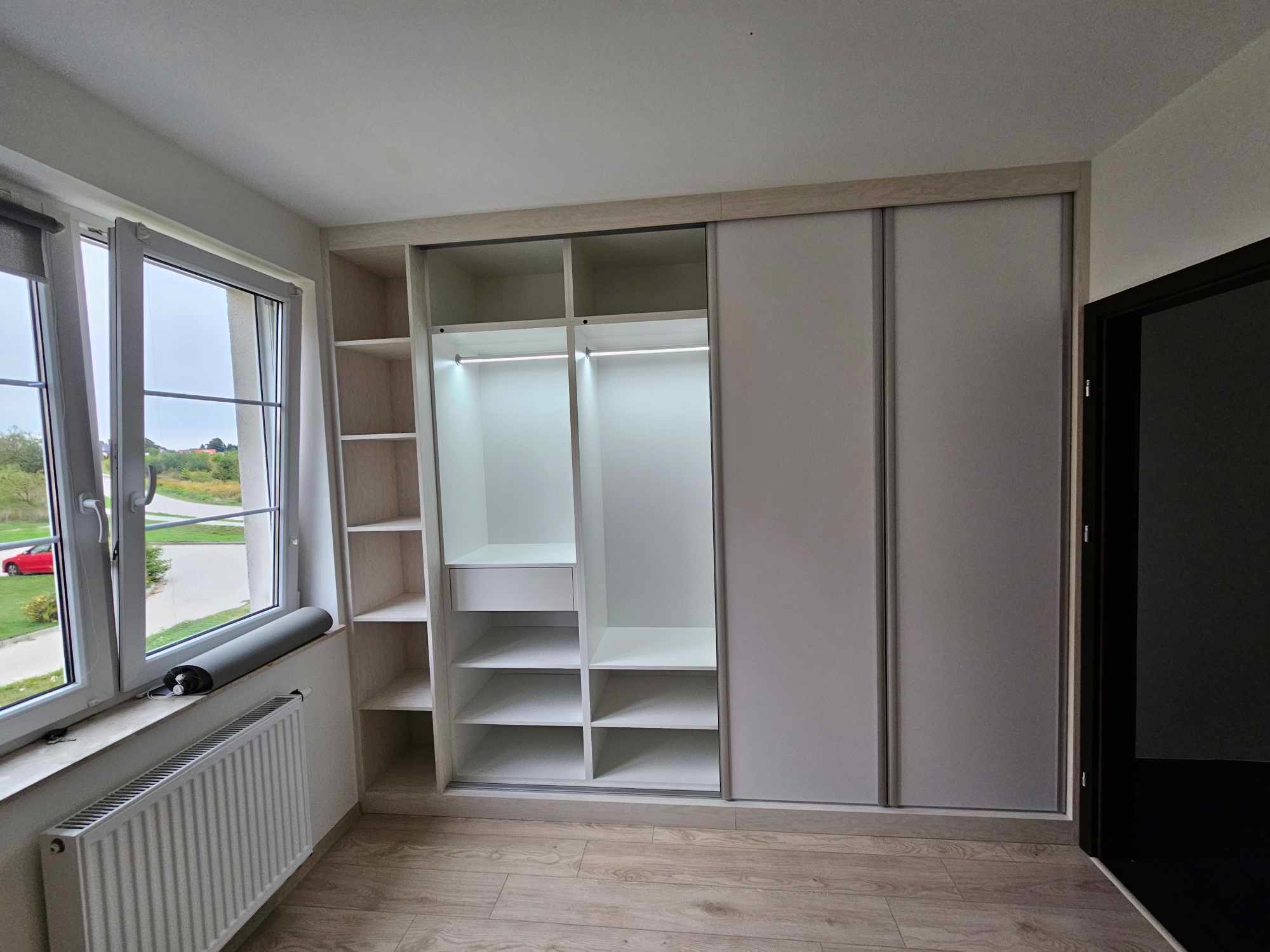 Nowoczesna szafa wnękowa z przesuwnymi drzwiami w jasnym pokoju, widok na okno z roletą i grzejnikiem, oświetlenie LED wewnątrz szafy.