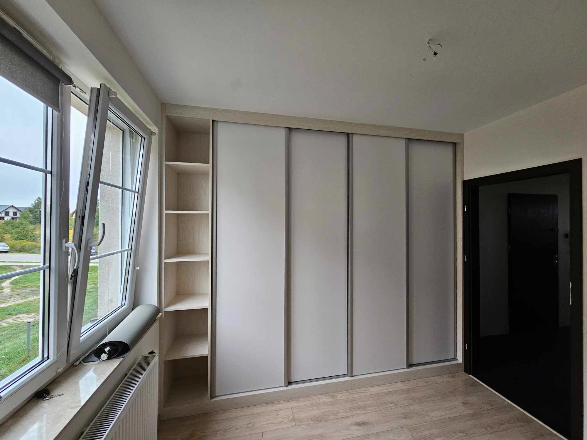 Wnętrze pokoju z dużą szafą wnękową z przesuwnymi, jasnoszarymi drzwiami, po lewej stronie regał z półkami, w tle okno z roletą i uchylone, widok na zabudowę jednorodzinną.