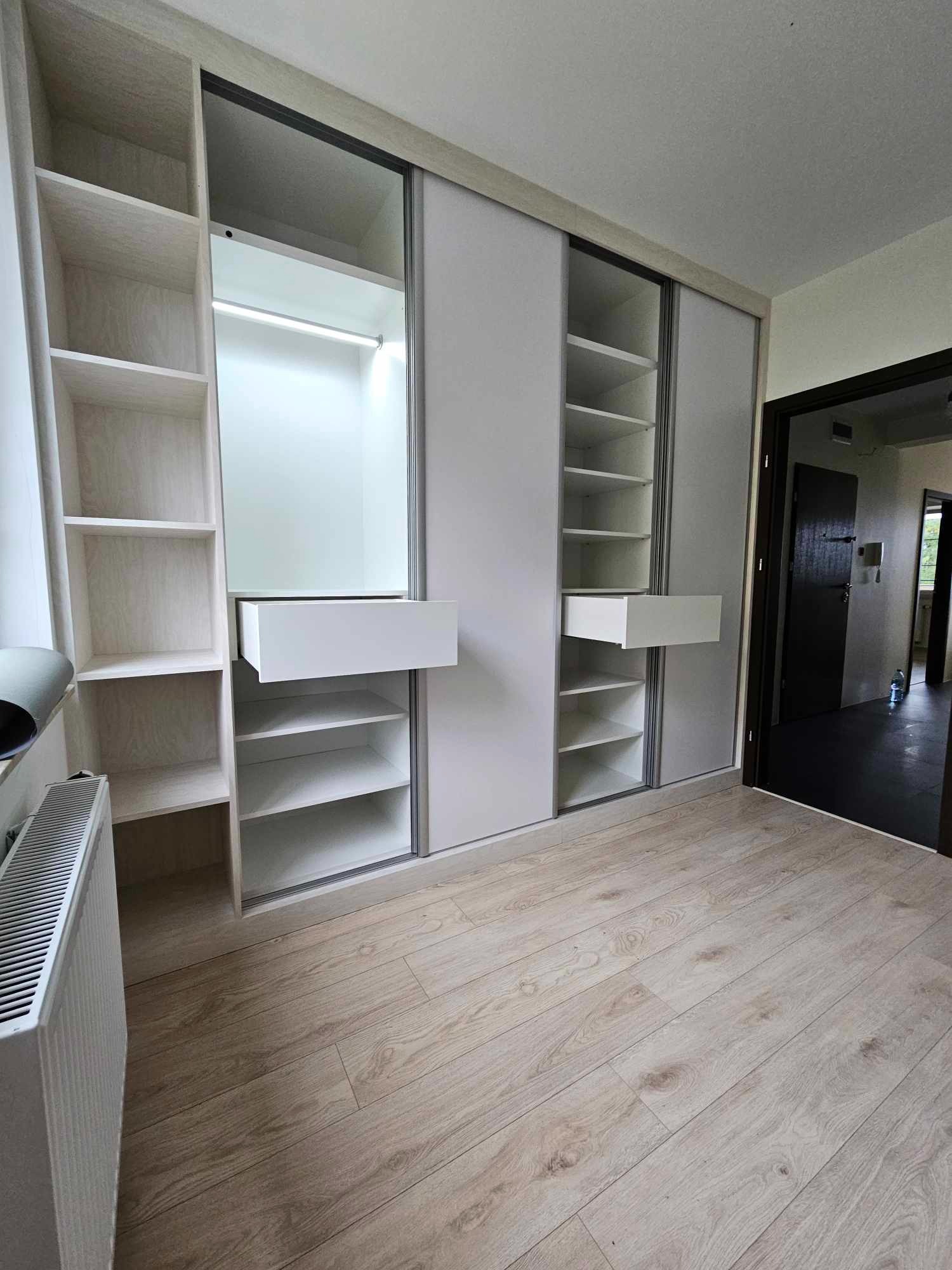 Jasna szafa wnękowa z przesuwnymi drzwiami i otwartymi półkami, z wysuniętą szufladą, oświetleniem wewnętrznym i drewnianą podłogą, widoczny grzejnik i drzwi do innego pomieszczenia.