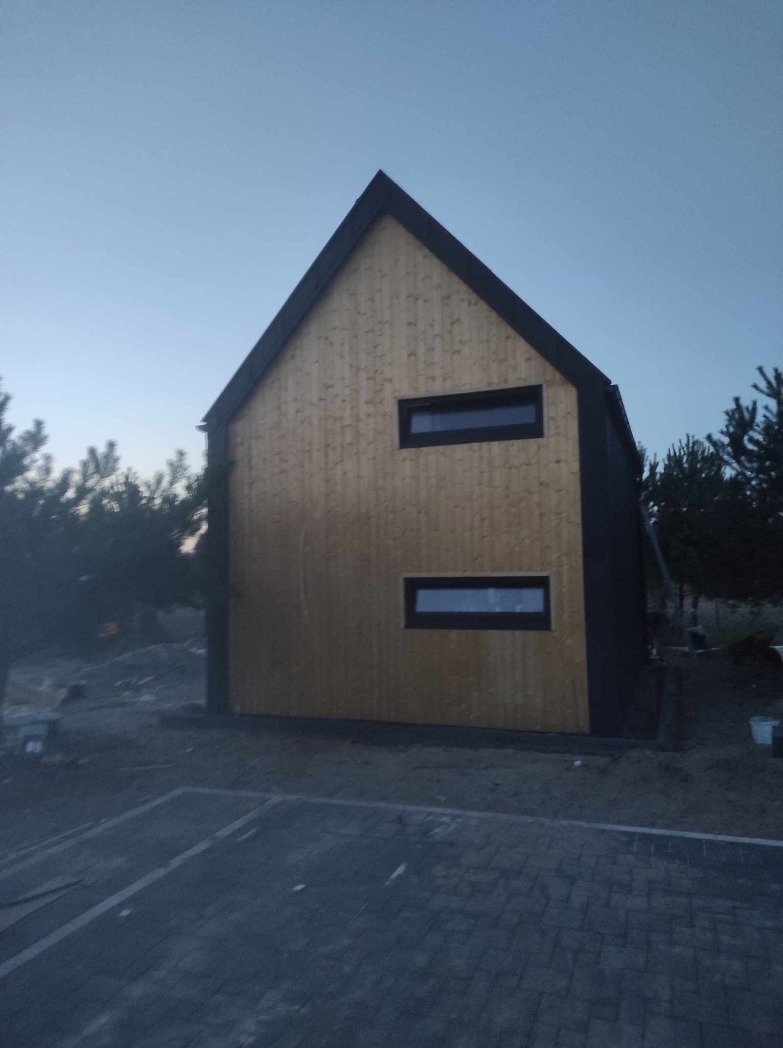 Nowoczesny dom z drewnianą elewacją i prostokątnymi oknami, widoczny o zmierzchu, z brukowanym podjazdem i drzewami w tle.