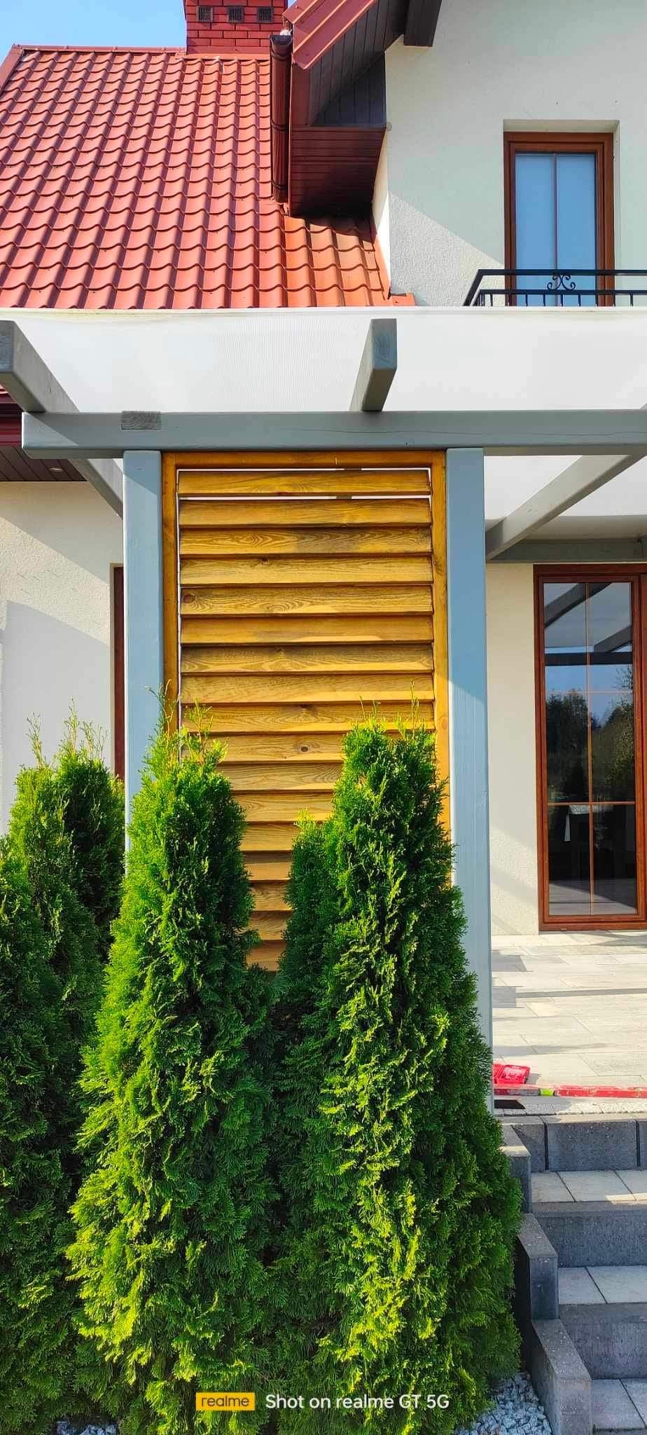 Drewniana żaluzja wbudowana w konstrukcję altany, osłaniająca od słońca, z widocznymi tujami w pierwszym planie i fragmentem domu w tle.