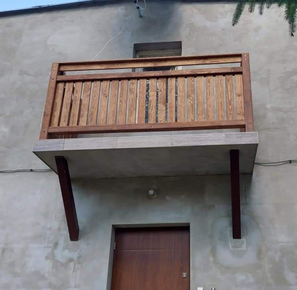 Mały, drewniany balkon z balustradą na betonowej płycie, wsparty na dwóch brązowych słupach, zamontowany nad drzwiami wejściowymi do budynku o szarej, surowej elewacji.
