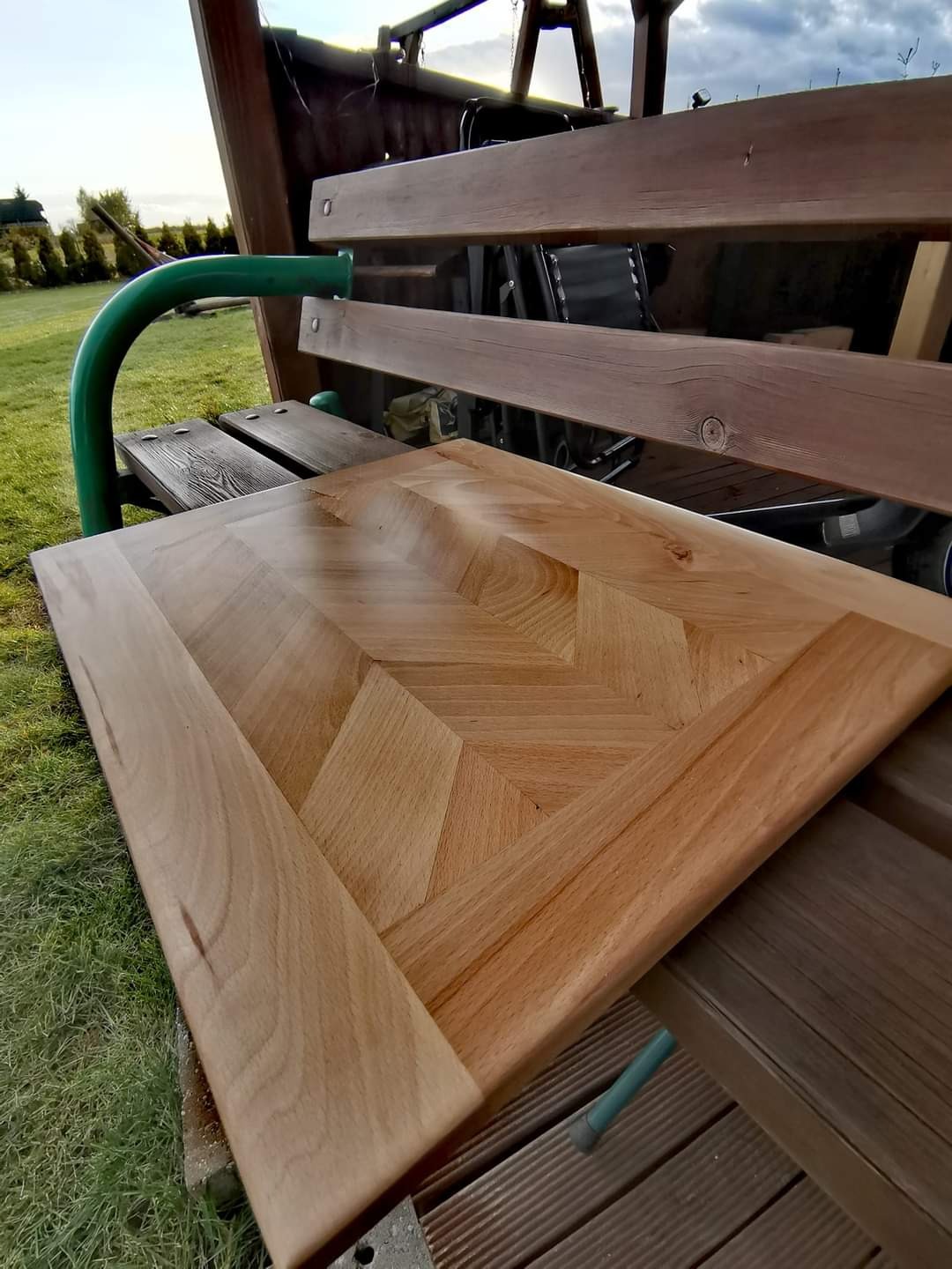 Blat stołu z drewna ułożonego w geometryczny wzór jodełki, oparty o drewnianą konstrukcję huśtawki ogrodowej, widoczne fragmenty trawnika i nieba.