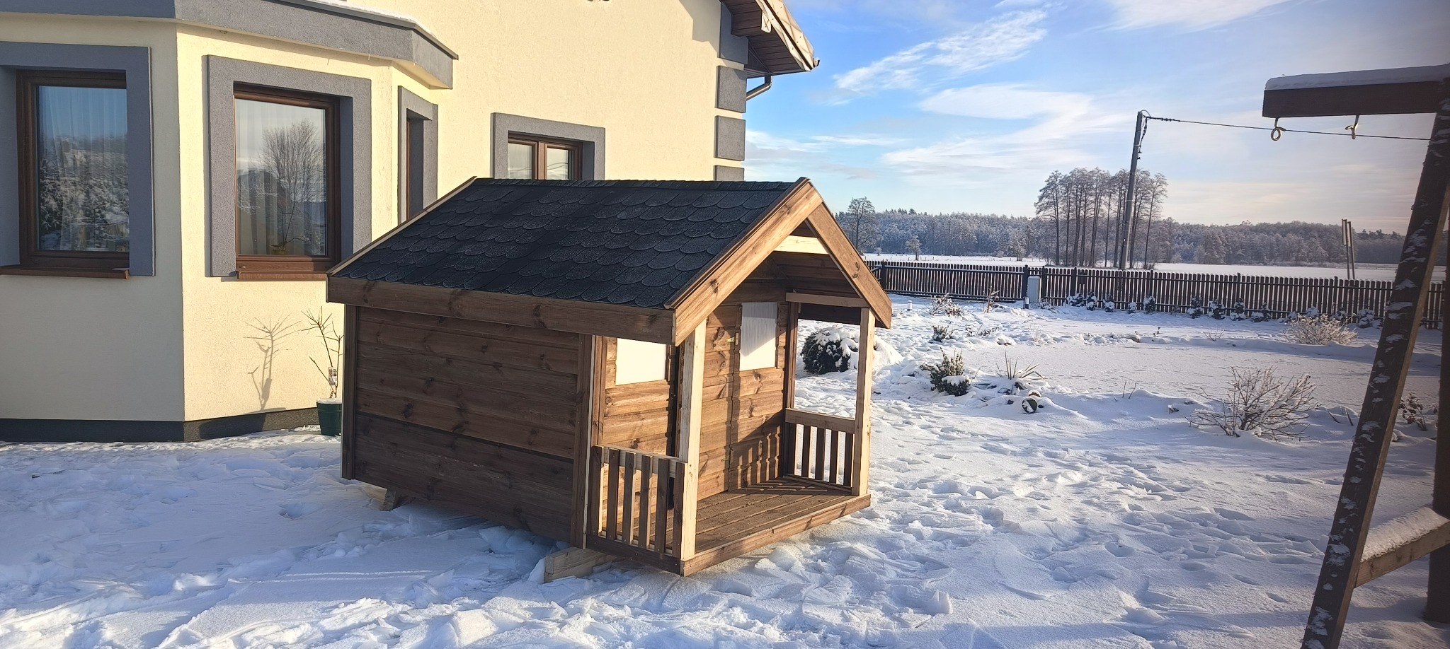 Drewniana konstrukcja, prawdopodobnie domek dla dzieci, z czarnym dachem bitumicznym, ustawiona na śniegu przed domem w słoneczny zimowy dzień.
