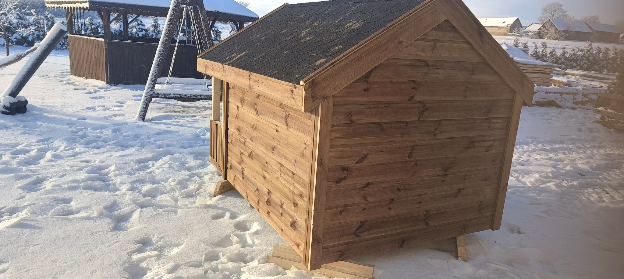 Mała, drewniana konstrukcja z dwuspadowym dachem pokrytym czarnym gontem bitumicznym, stojąca na drewnianych legarach w zimowym otoczeniu, z widoczną altaną w tle.