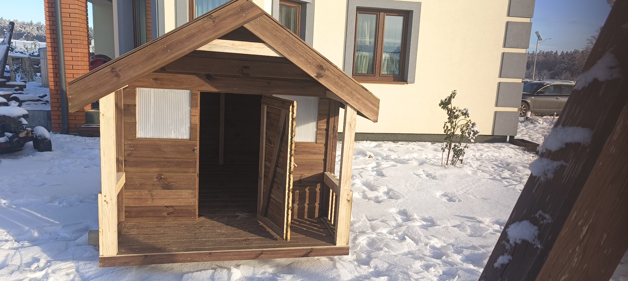 Drewniana altana dla dzieci z otwartymi drzwiami, częściowo pokryta śniegiem, na tle zimowego ogrodu i domu.