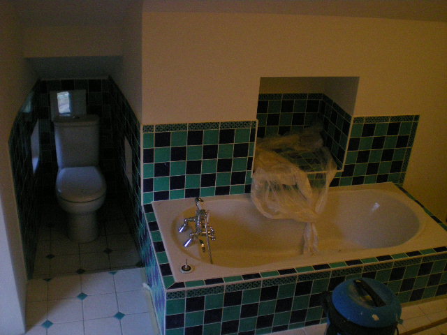 Łazienka w trakcie remontu: widoczna wanna zabezpieczona folią, nowa glazura w odcieniach zieleni i granatu, oraz toaleta w małej wnęce.