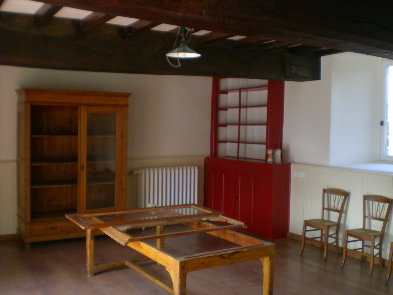 Pusta przestrzeń z drewnianą szafą z przeszklonymi drzwiami, czerwonym regałem, grzejnikiem i rozłożonym drewnianym stołem z szklanym blatem w trakcie renowacji, pod sufitem widoczne drewniane...