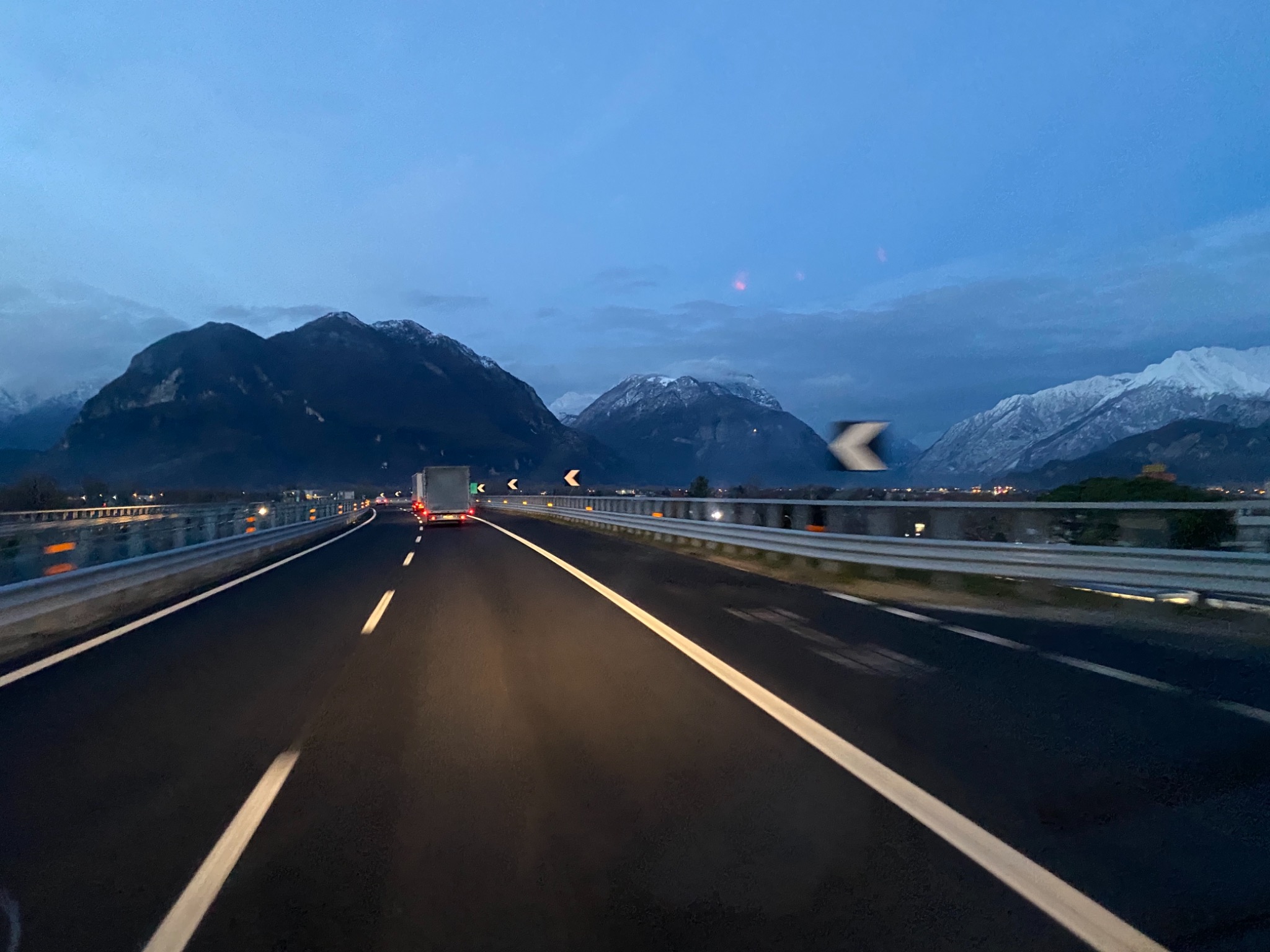 Jazda autostradą o zmierzchu; w oddali widoczne góry z ośnieżonymi szczytami i jadąca ciężarówka.