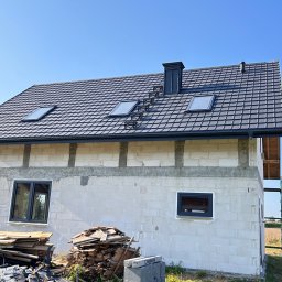 DACH-SYSTEM - Dom w trakcie budowy z nowym, ciemnoszarym dachem z trzema oknami dachowymi i kominem z drabinką kominiarską, ściany z białego bloczka częściowo otynkowane, widoczne rusztowanie i sterta odpadów...