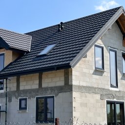 DACH-SYSTEM - Dom w trakcie budowy z nowym, ciemnoszarym dachem z dachówki modułowej, oknem dachowym i kominkami wentylacyjnymi, widoczny fragment rusztowania i reklama dekarza na ścianie.