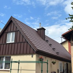 DACH-SYSTEM - Dom z nowym, brązowym dachem z blachy trapezowej, kominem i rynnami, numer posesji '8', kwiaty w wiszących donicach, częściowo widoczne rusztowanie.