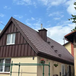 Dom z nowym, brązowym dachem z blachy trapezowej, kominem i rynnami, numer posesji '8', kwiaty w wiszących donicach, częściowo widoczne rusztowanie.